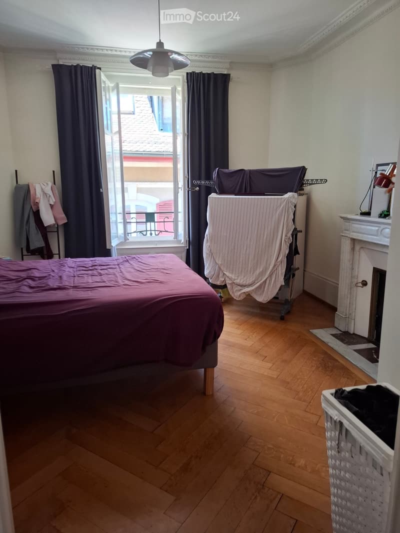Bedroom with wooden flooring, a bed with purple bedding, a washing machine, a fireplace, a lamp, and a clothing rack with clothes.
