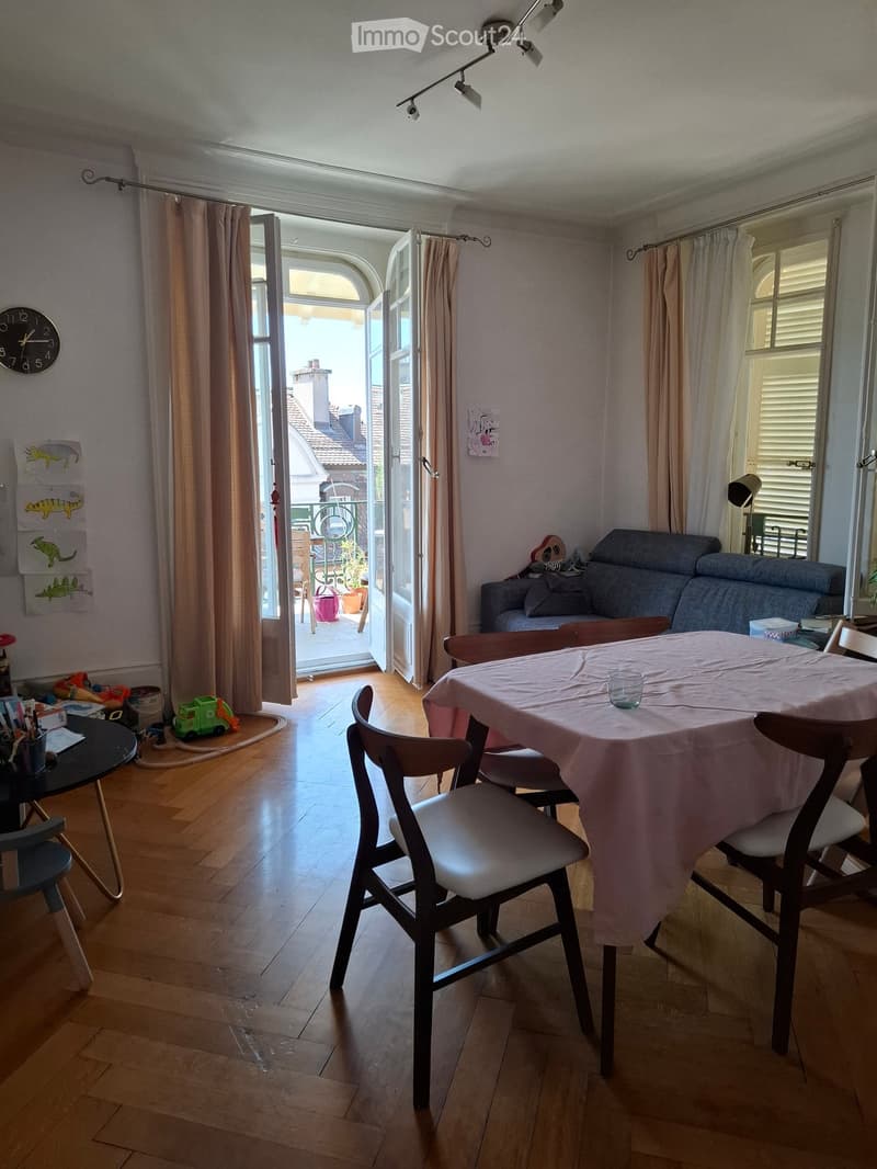 Bright living and dining area with wooden flooring, large open sliding doors leading to a balcony, a sofa, a dining table with chairs, a clock on the wall, and a guitar leaning against the wall.