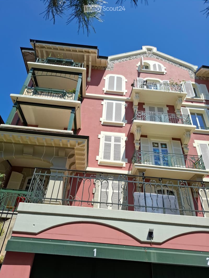 Multi-story building with red and cream facade, multiple balconies with metal railings, ornate architectural details, and shutters on windows.