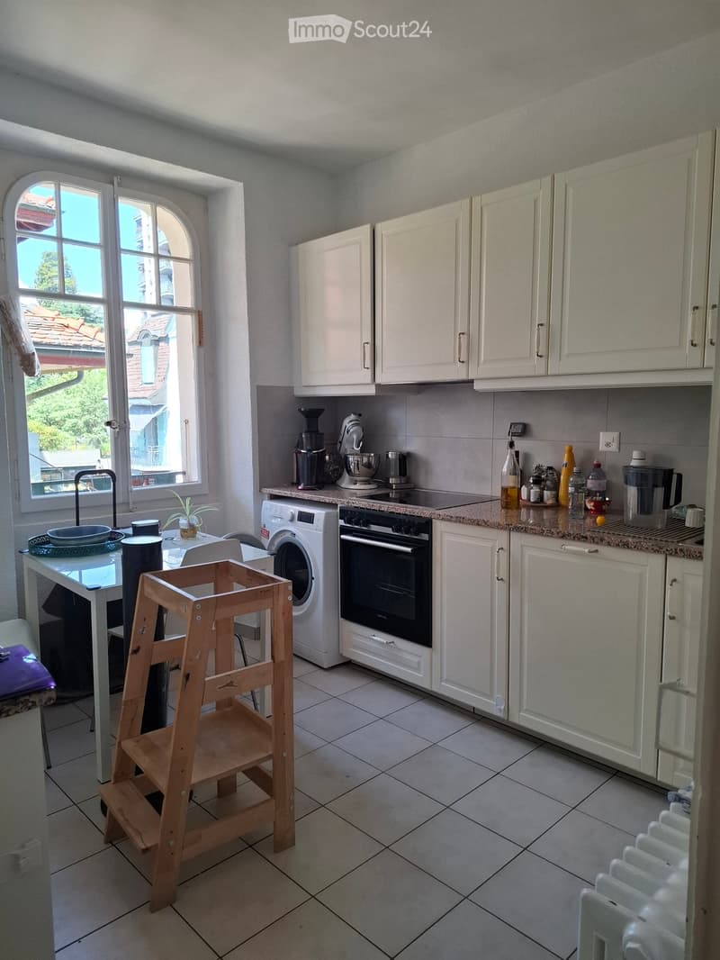 A modern kitchen with white cabinets, a tiled floor, and various appliances including an oven, washing machine, and coffee maker. There is a small wooden stool, a table, and a window with a view of the outside.