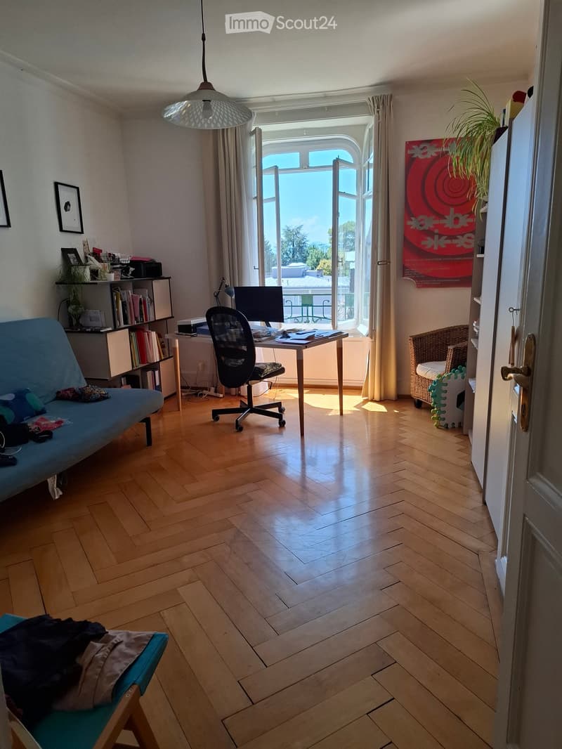 Bright living room with wooden flooring, large windows, and a balcony view. Features a blue sofa, a desk with a computer, a bookshelf, and a hanging lamp.