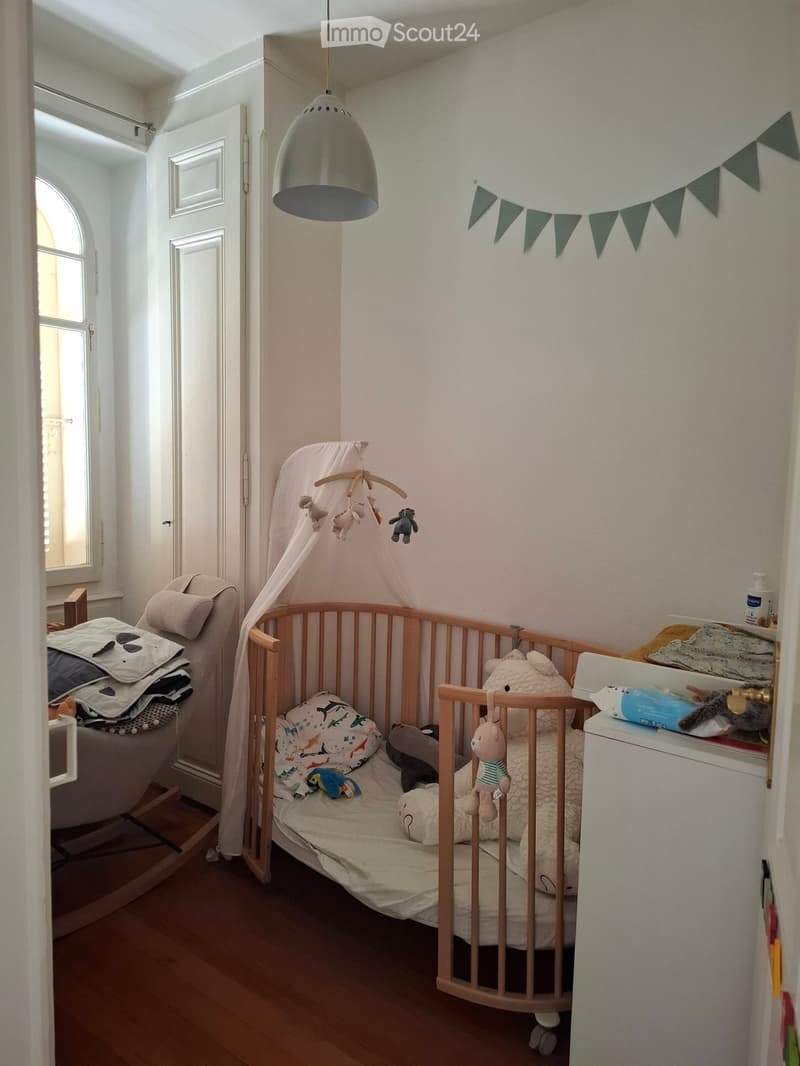 A well-lit children's bedroom with a wooden crib, a sofa, toys, and a hanging mobile above the crib.