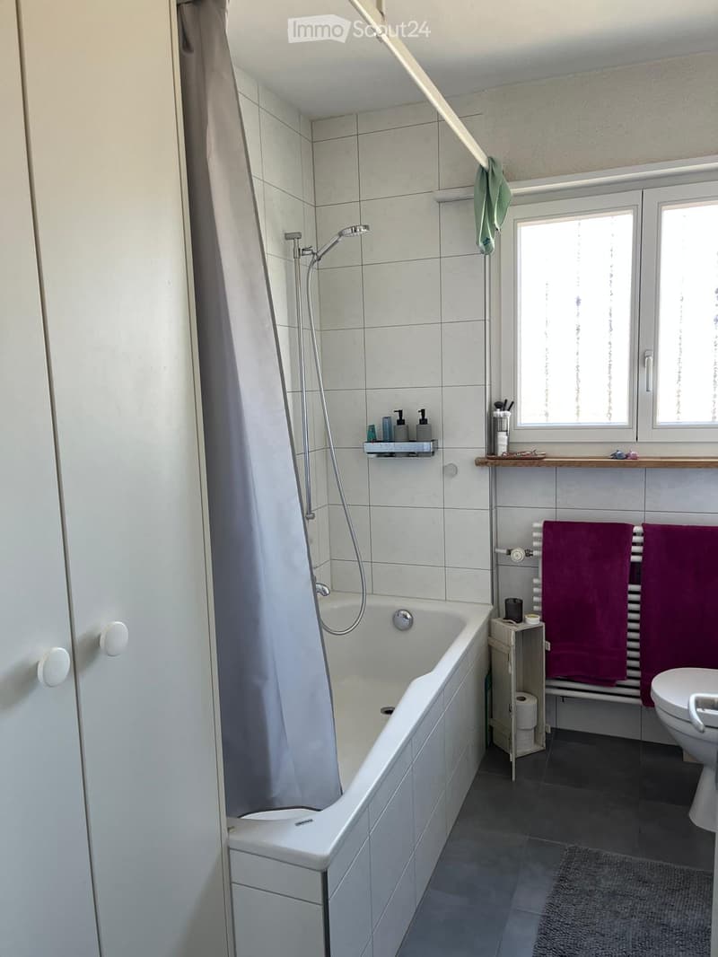 Modern bathroom with a white bathtub, shower, toilet, towel rack, and small cabinet. The room features tiled walls and a window providing natural light.