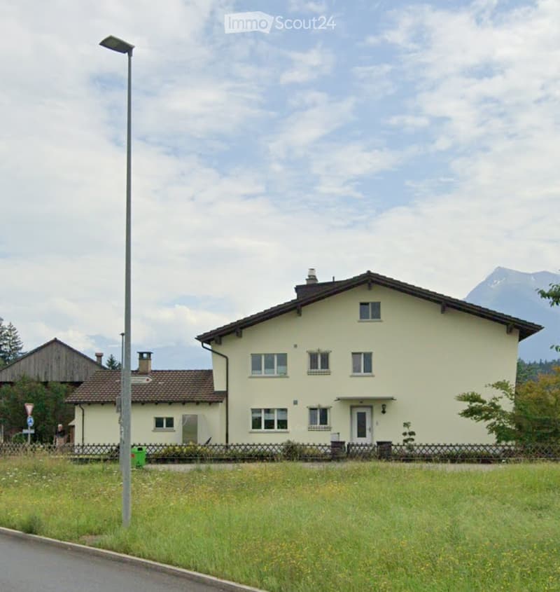 Two-story house with beige walls, multiple windows, and a tiled roof, situated in a grassy area with a fence and a streetlight nearby.