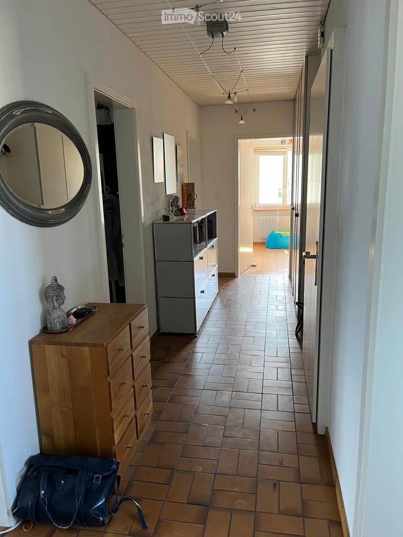 A narrow hallway with tiled flooring, white walls, and a wooden dresser on the left side. A round mirror is mounted on the wall, and a small table with a decorative item sits near the dresser. The hallway leads to an open area with a visible sofa and a wi