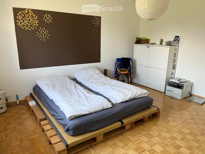 A modern bedroom with a wooden floor, a bed with white bedding, a white wardrobe, a chair with a jacket, and a printer on a shelf.