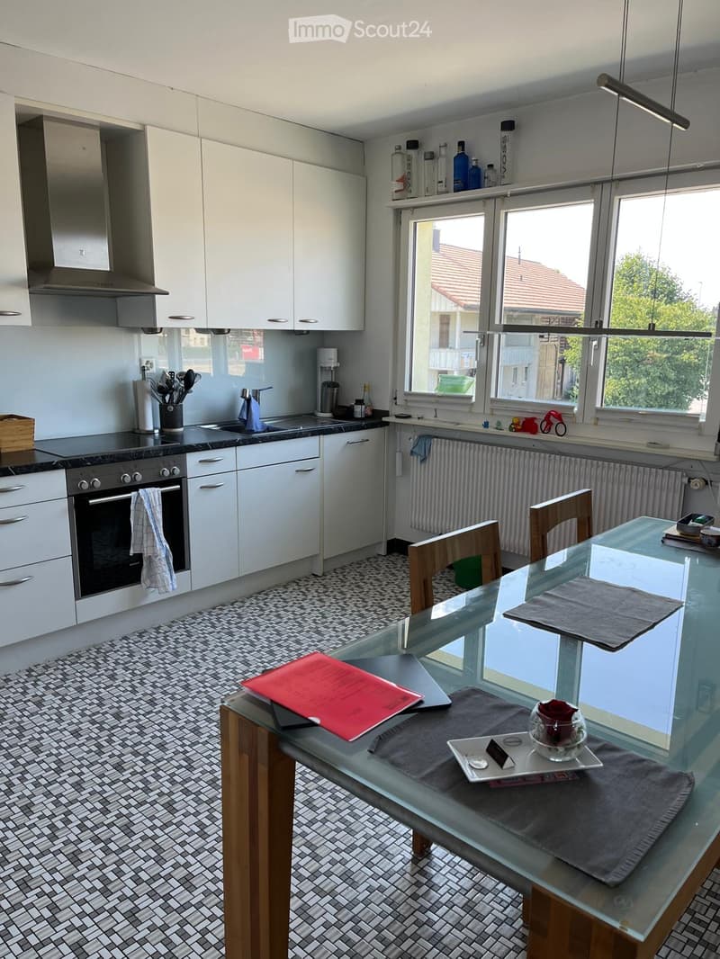 Modern kitchen with white cabinets, black countertop, glass dining table, and wooden chairs. Features include an oven, sink, and various kitchen utensils.