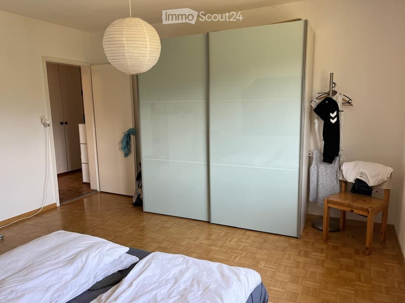 Bedroom with wooden flooring, a bed with white sheets, a large wardrobe, a small wooden chair, and a coat rack with clothes.