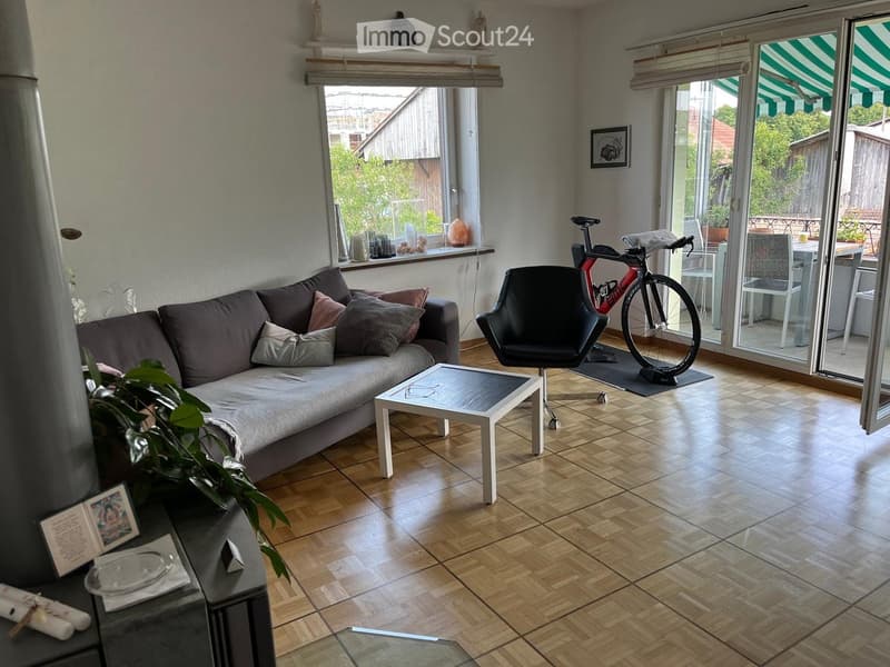 Modern living room with wooden flooring, large windows, a grey sectional sofa with throw pillows, a white coffee table, a black chair, a bicycle, and a small table with chairs near the balcony door.