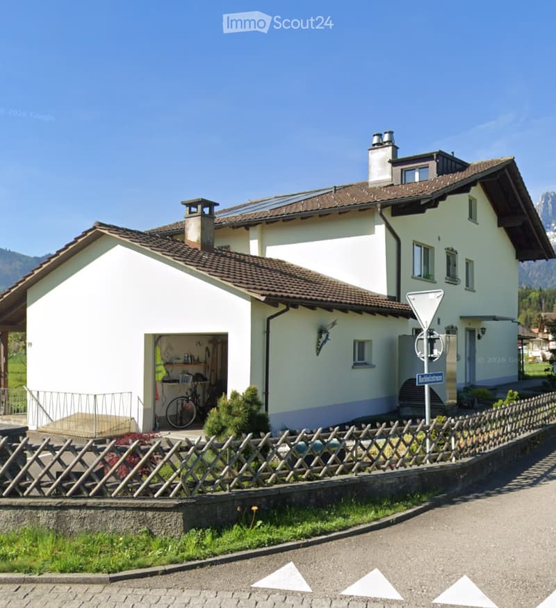 Two-story house with white walls, brown tiled roof, visible garage, and small garden area.