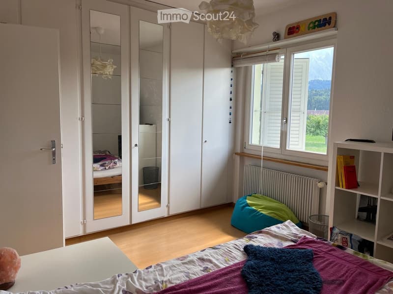 Bright bedroom with wooden flooring, large window with view, bed with colorful bedding, mirror wardrobe, shelf with books, beanie chair, and a small table.