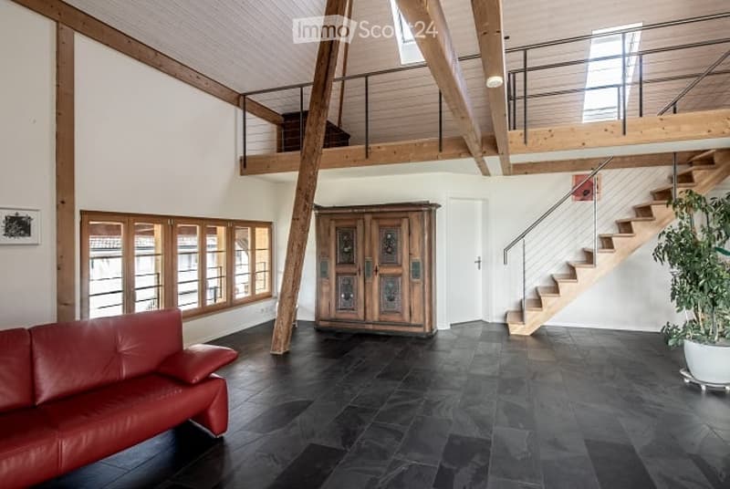 Modern living room with dark tiled flooring, exposed wooden beams, large windows, red leather sofa, wooden cabinet, staircase leading to an upper level, and a potted plant near the stairs.
