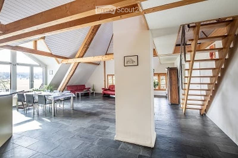 Open-plan living and dining area with exposed wooden beams, modern furniture including a red sofa, dining table with chairs, and a small stool.