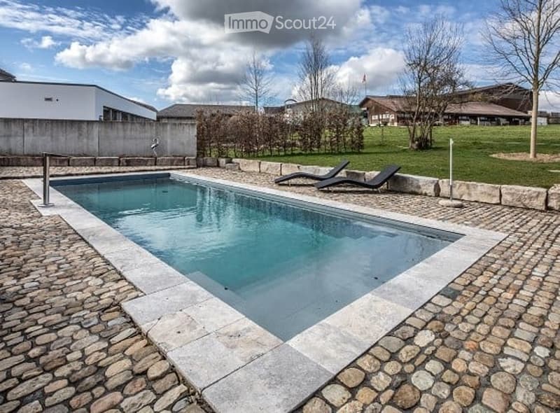 Outdoor swimming pool surrounded by stone tiles, with two lounge chairs placed nearby, surrounded by a grassy area and trees.