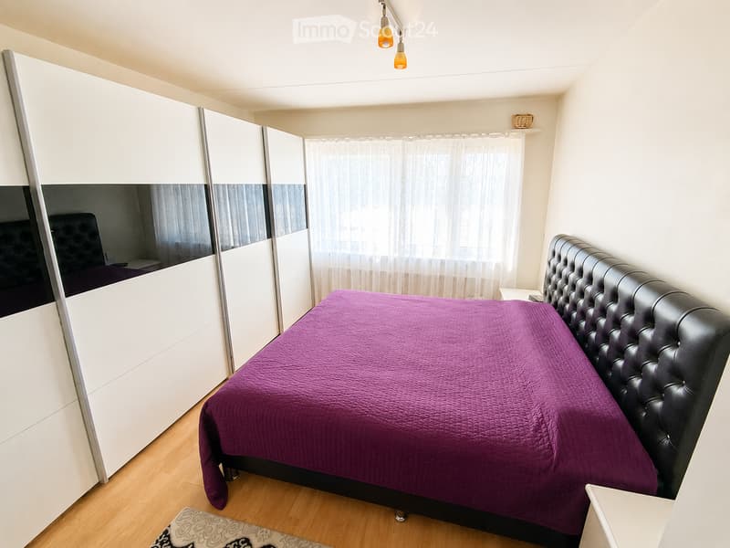 Modern bedroom with a large bed covered in a purple duvet, a black leather headboard, a white sliding wardrobe, and a small nightstand.