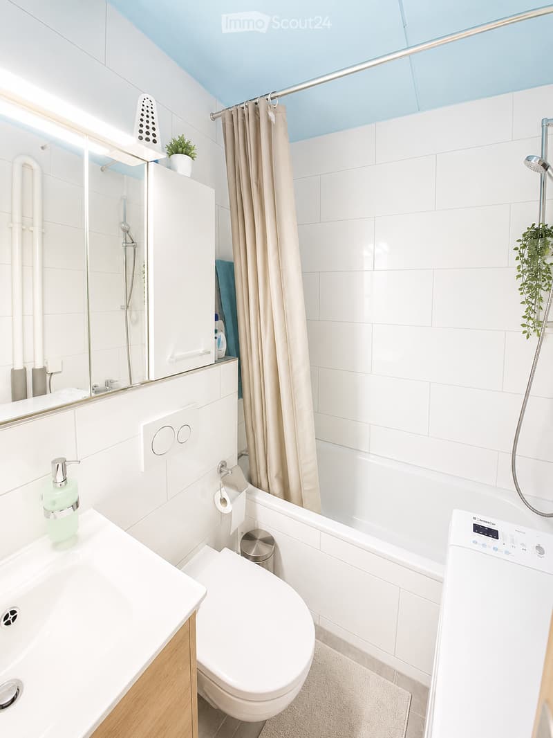 Modern bathroom with white tiles, a bathtub, toilet, sink, and a shower area with a curtain. Includes a mirror, cabinet, and a small plant on the windowsill.