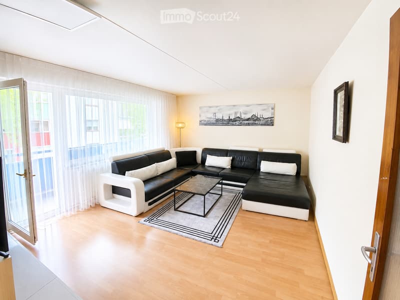 Modern living room with a black and white sectional sofa, a glass coffee table, a floor lamp, and a large picture frame on the wall. The room features wooden flooring and large windows with sheer curtains.