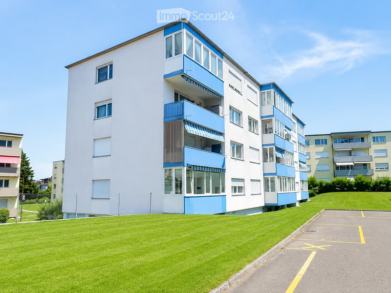 A modern multi-story residential building with white and blue accents, featuring balconies and large windows, surrounded by a well-maintained lawn and paved pathways.