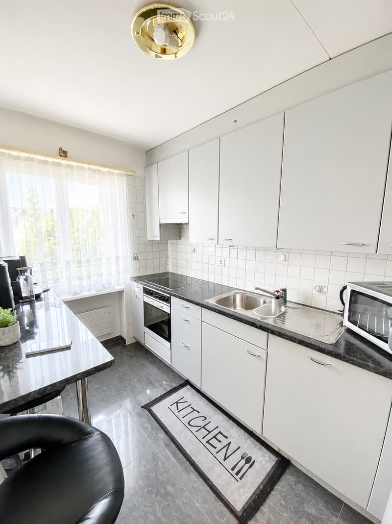 Modern kitchen with white cabinets, black countertops, stainless steel sink, oven, microwave, and a small table with a chair. A decorative rug with the word "KITCHEN" is on the floor.