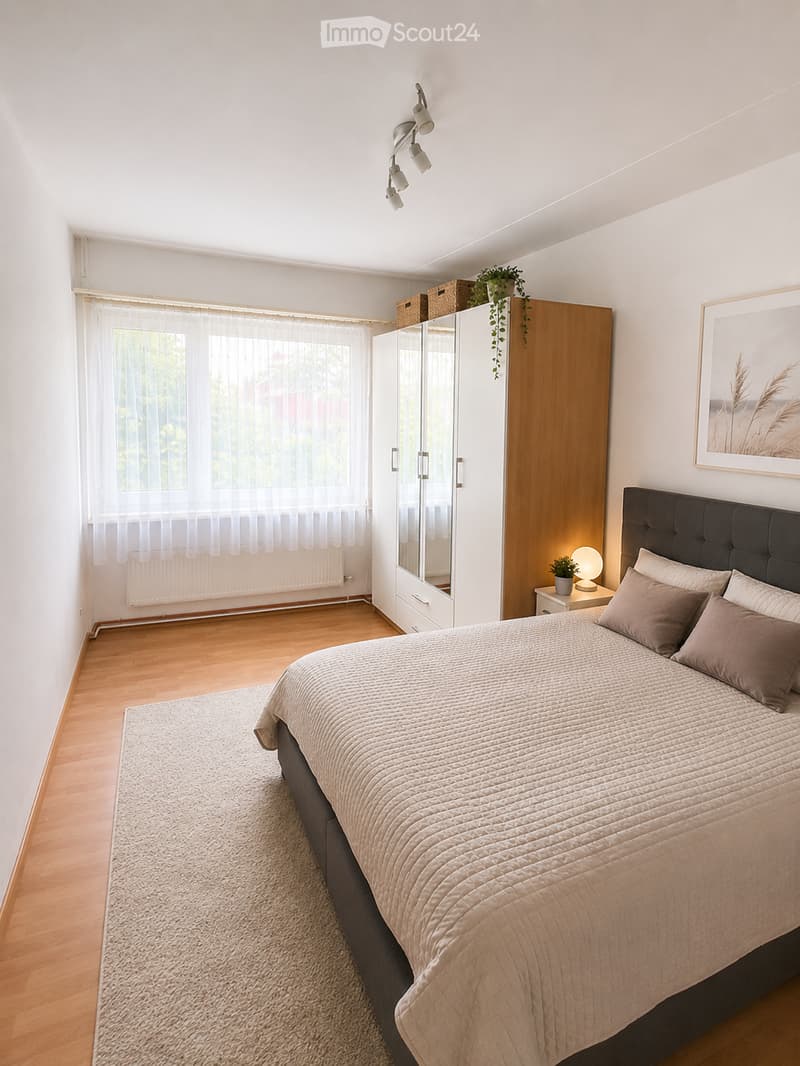 Modern bedroom with a large bed, nightstand, lamp, wardrobe with mirror, and a potted plant on the nightstand. The room features wooden flooring, large windows with sheer curtains, and a cozy rug.