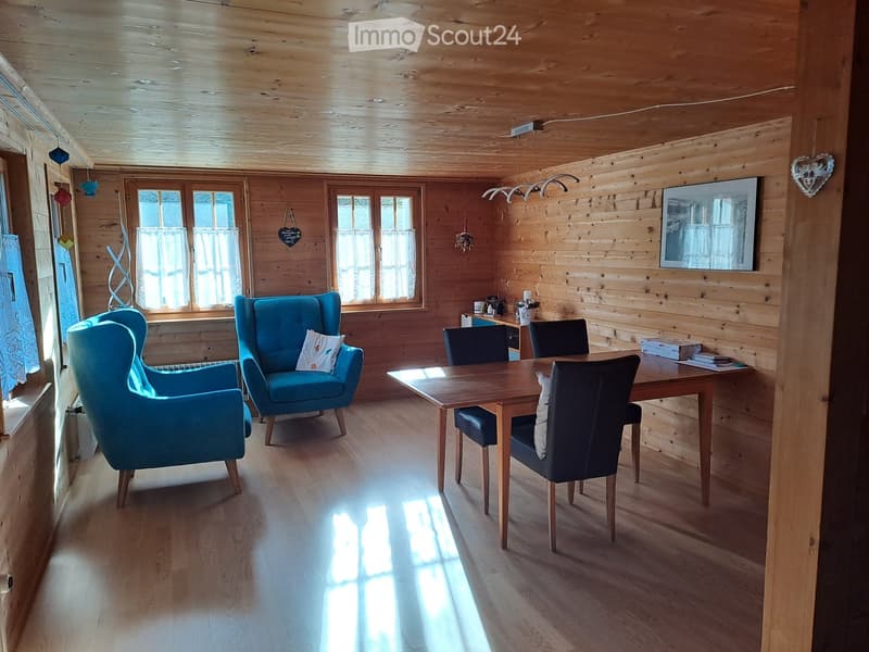 Cozy living room with wooden walls and ceiling, large windows, blue armchairs, wooden dining table with chairs, and a mirror on the wall.