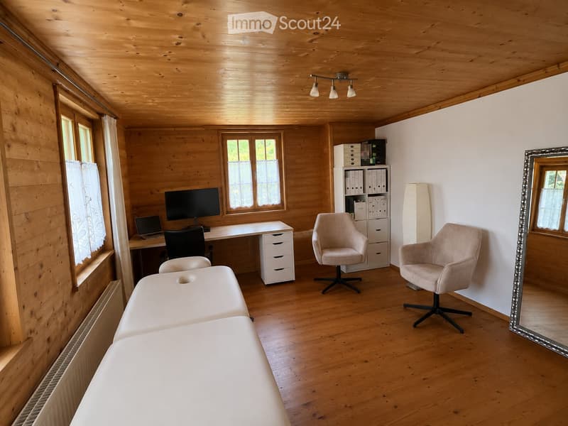 A well-lit room with wooden walls and ceiling, featuring a desk with a laptop and screen, two chairs, a large mirror, and a storage unit with files.