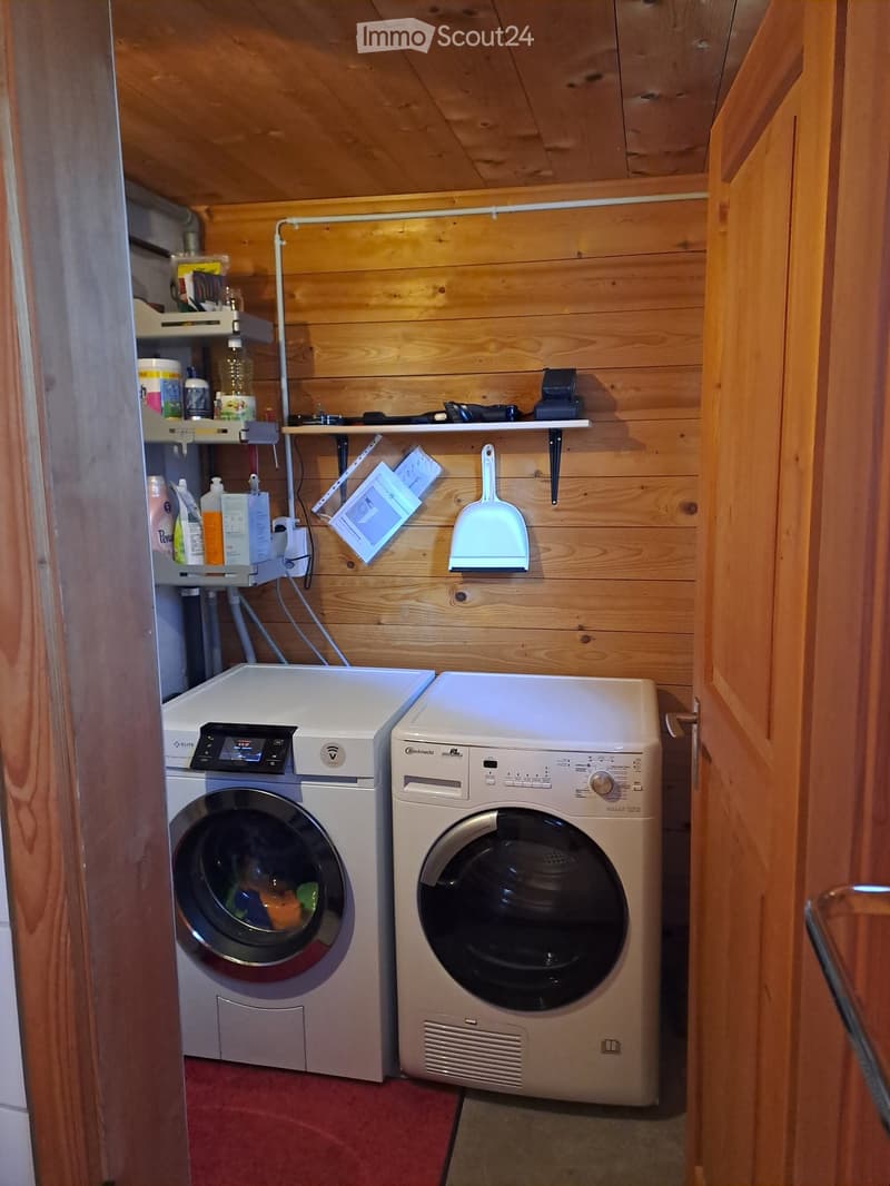 A small, wooden-paneled laundry area with a washing machine and dryer side by side, shelves holding cleaning supplies, and a broom hanging on the wall.