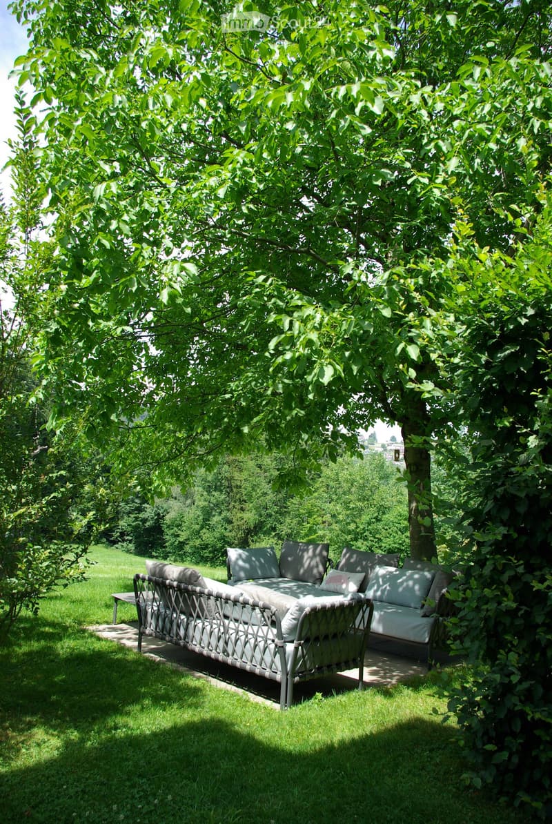 A garden area with a large tree providing shade, featuring a white outdoor sofa with cushions and a small table.