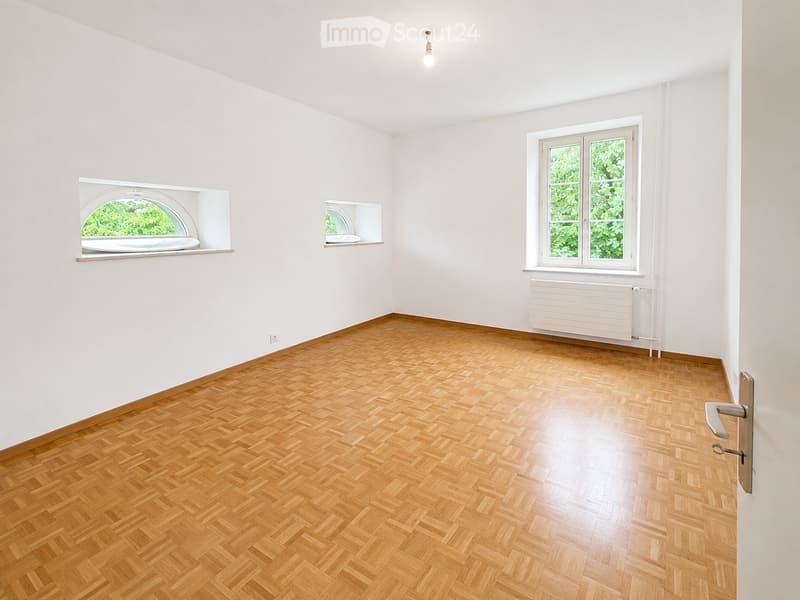 Empty room with wooden herringbone flooring, white walls, and large windows with green views. Features include a built-in shelf and radiator.