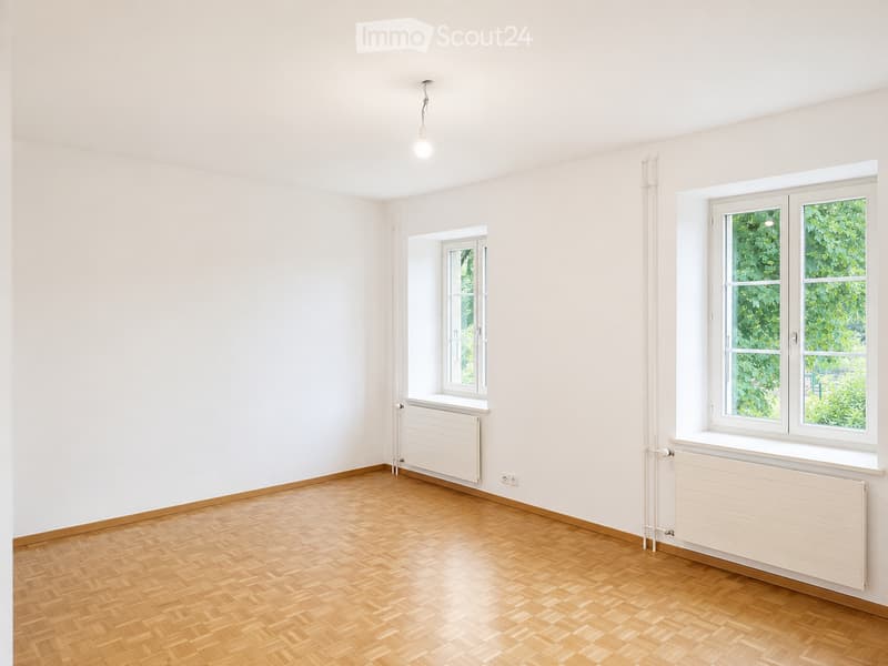 Empty room with light-colored walls, wooden herringbone flooring, and two windows with white frames. Radiators are present beneath each window.