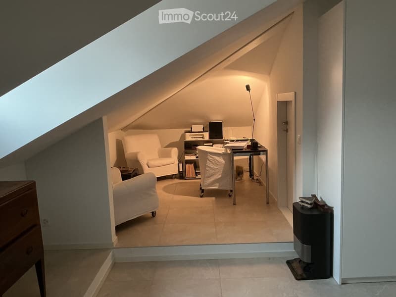 Cozy attic room with slanted ceilings, white furniture including a sofa, chairs, a small table, and a lamp, with a bookshelf and printer visible.