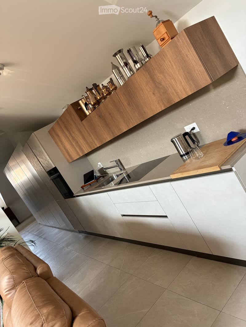 Modern kitchen with wooden cabinets, stainless steel appliances, and a sleek sink. A leather sofa is visible in the foreground.
