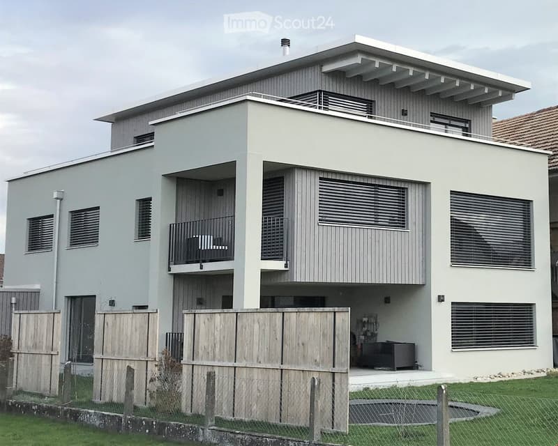 Modern two-story house with light grey exterior, large windows, balconies, and a fenced yard.