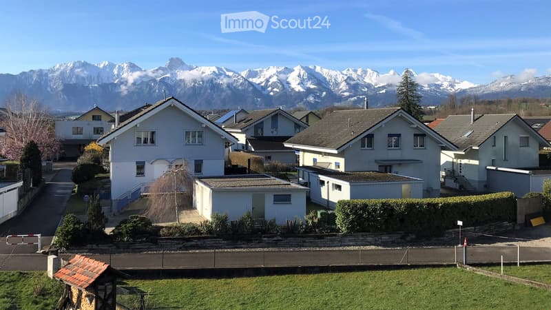 Residential neighborhood with multiple houses, surrounded by greenery and mountains in the background.