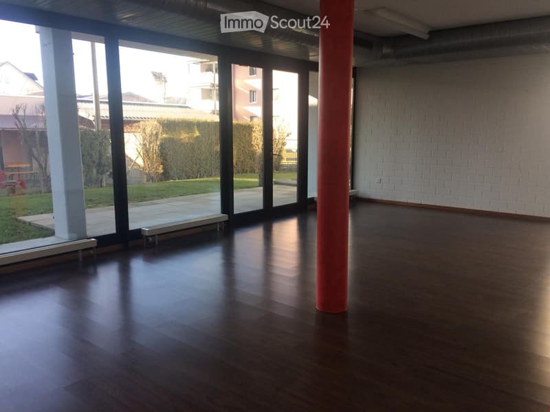 Empty room with dark wooden flooring, white brick wall, large glass windows, and a red column. No furniture or equipment present.