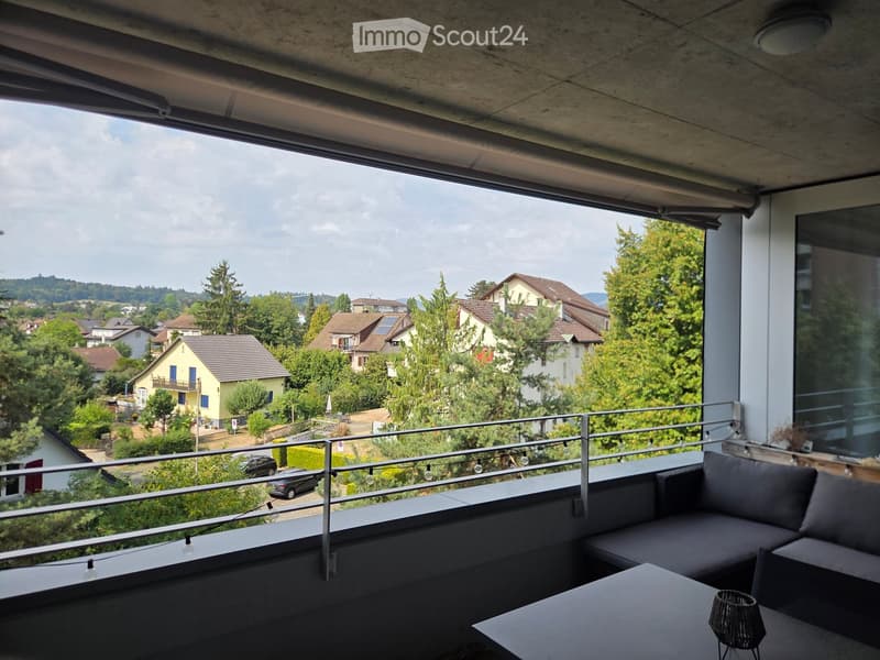 Balcony with modern furniture, including a sofa, table, and lamp, overlooking a scenic residential area with houses and greenery.