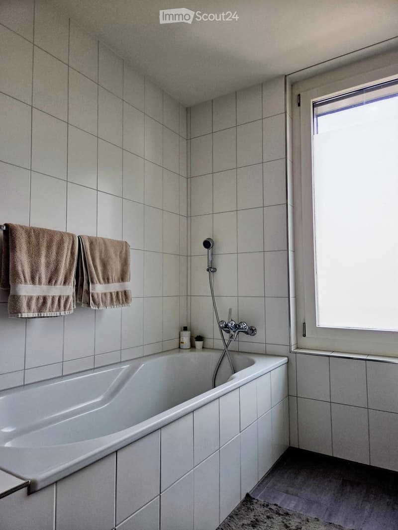 Bathroom with a white bathtub, tiled walls, a window, and two towels hanging on a rack.