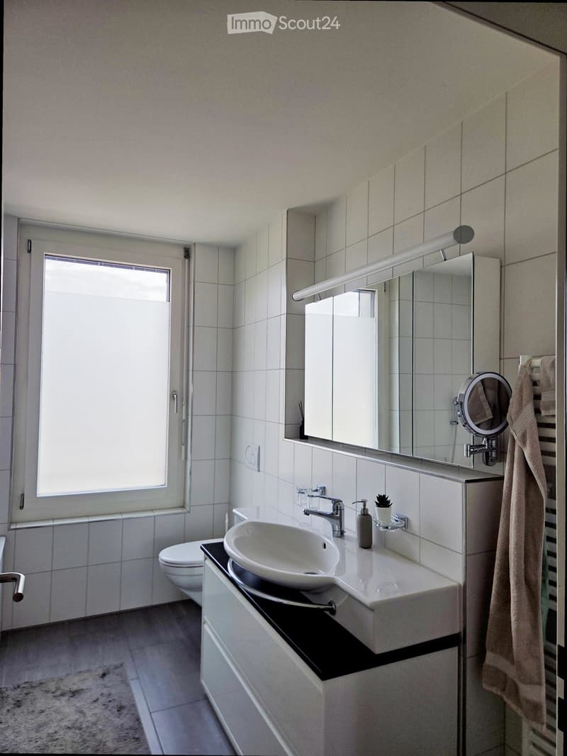 Modern bathroom with white tiles, a large sink with a modern faucet, a mirror with built-in lighting, a toilet, and a towel hanging on the side.