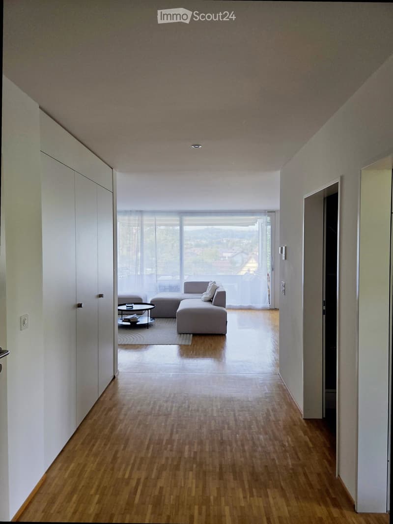 Modern hallway with wooden flooring, white walls, and recessed lighting. A large window at the end provides natural light and a view of the living area, which includes a grey sectional sofa, coffee table, and side table.