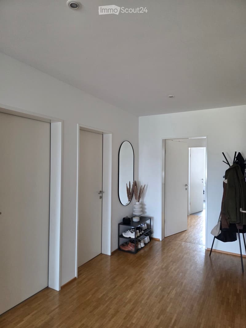 Hallway with wooden flooring, white walls, a mirror, a vase with dried plants, a shoe rack, and a coat rack with jackets.
