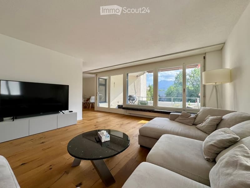 Modern living room with large windows offering a scenic view, featuring a grey sectional sofa, a round glass coffee table, a flat-screen TV on a white stand, and a floor lamp.