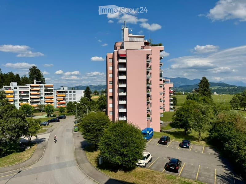 Aerial view of a multi-story residential building with pink facade and balconies, surrounded by greenery and a parking lot with several cars.