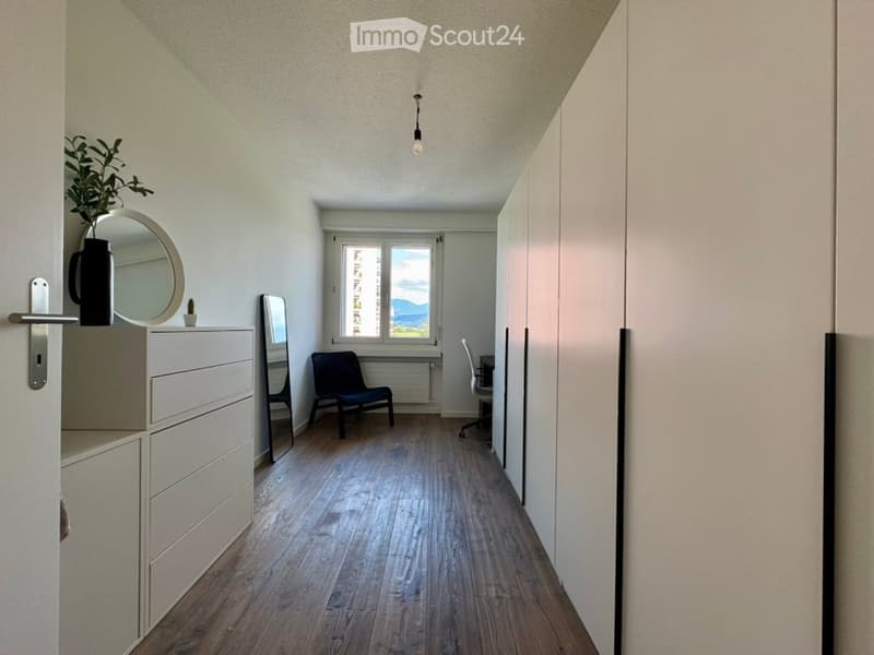 A narrow, well-lit hallway with wooden flooring, a small black chair, a mirror, a drawer unit, and a desk with a chair. A potted plant is placed on the drawer unit, and a round mirror hangs on the wall.