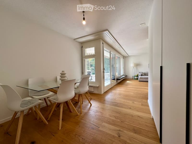 Bright, open-plan dining and living area with wooden flooring, large glass windows, a modern dining table with chairs, and a sofa in the background.