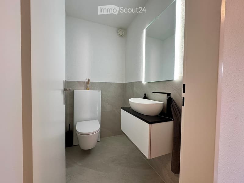 Modern bathroom with a white toilet, a sink with a modern faucet, a large mirror, and a white cabinet. The walls are tiled in a neutral color scheme, and the floor is covered with light gray tiles.