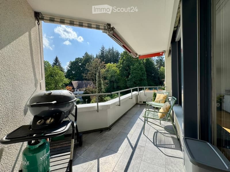 Balcony with a barbecue, two chairs, and a small table, overlooking a green, tree-filled landscape.