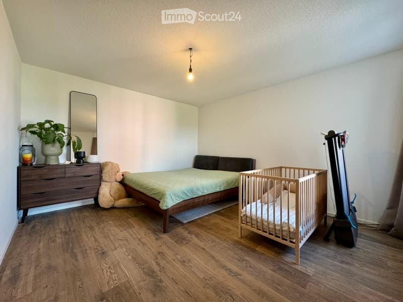 Cozy bedroom with a wooden bed, crib, dresser, mirror, plant, teddy bear, and a hanging lamp.