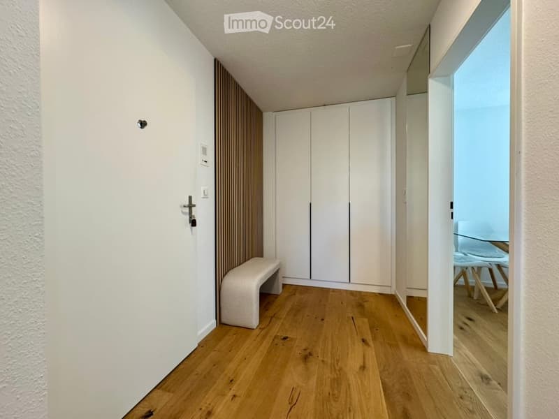 A modern hallway with light wooden flooring, white walls, and a built-in white closet. A small white bench is placed against the wall, and a glass door leads to another room with a visible table and chairs.