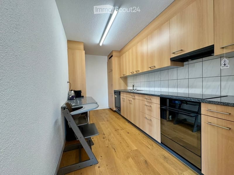 Modern kitchen with light wooden cabinets, granite countertops, and stainless steel appliances including a dishwasher and oven. A small table with a stool is visible in the corner.