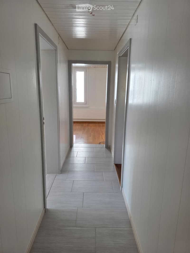 Clean, modern hallway with tiled flooring and white walls, featuring several doors and a visible room at the end with wooden flooring.